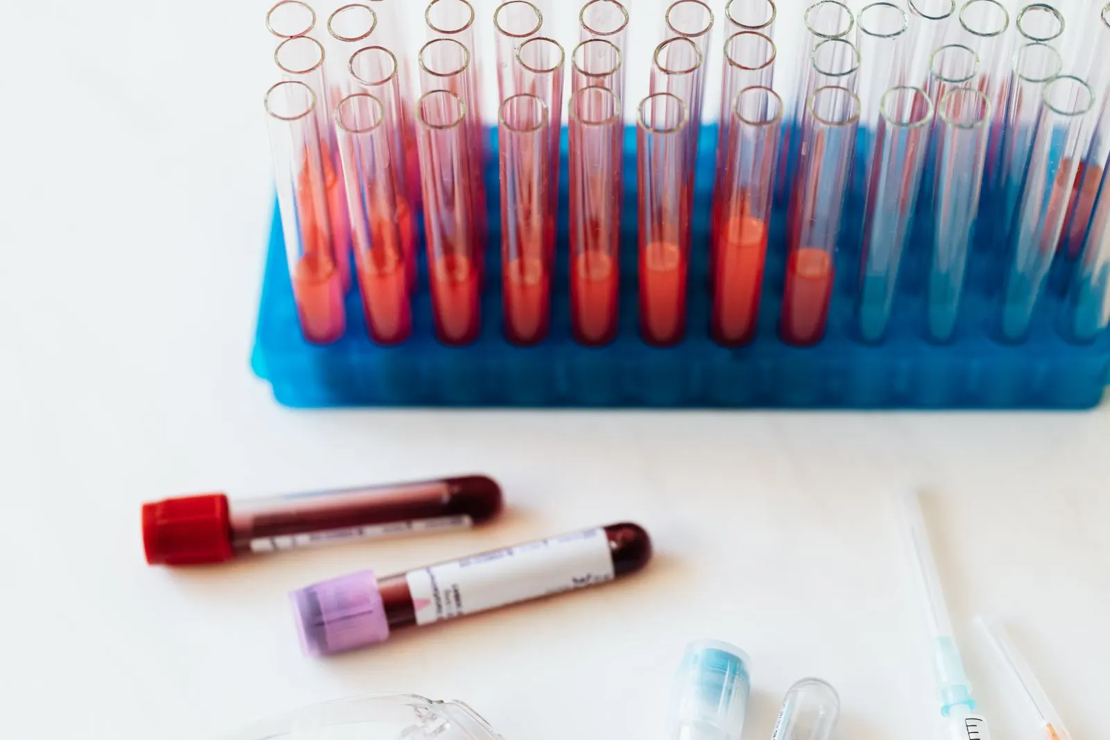 Probówki z próbkami krwi w niebieskim statywie i dwie zakręcone probówki leżące na białym blacie laboratorium.